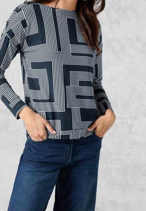 Femme les mains dans les poches, vêtue d'un haut à manches longues à motif géométrique noir et blanc et d'un jean bleu foncé sur un fond gris.