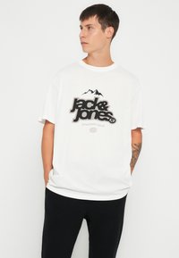 Camiseta de algodón blanca con un logo en negro y gris que presenta un gráfico de montaña, cuello redondo, corte holgado y mangas cortas. Combinada con pantalones negros.
