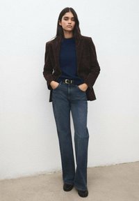 Rjav srajčni blazer iz korduroja, temnomoderen pulover, temno modre široke kavbojke z visokim pasom, črni čevlji; model stoji z rokami v žepih pred svetlim zidom.