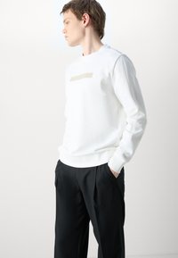 Felpa bianca con un leggero logo dorato sul petto, abbinata a pantaloni neri plissettati. Tessuto morbido, vestibilità rilassata.