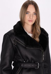 Veste en cuir noir avec un col en fourrure noire, des poches zippées et une taille ceinturée avec des accents en métal. La surface texturée ajoute de la profondeur.