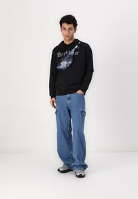 Hanorac negru cu logo-ul "Barbour", blugi largi de culoare albastru deschis și o geantă de umăr cu model de culoare albastră închisă. Adidași gri completează ținuta.