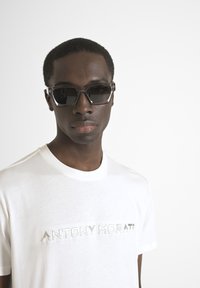 Gafas de sol con un marco rectangular grueso y tintado en gris. Combinadas con una camiseta blanca de algodón con un logo reflectante. Fondo sencillo.