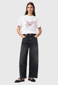 Λευκό μπλουζάκι με κόκκινο γραφικό σχέδιο, συνδυασμένο με μαύρο τζιν ψηλόμεσο παντελόνι με φθαρμένο τελείωμα και μαύρα επίπεδα παπούτσια.