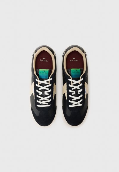 Black sneakers with suede and leather material, white laces, cream accents, and a colored label showcasing “PS Paul Smith” inside.