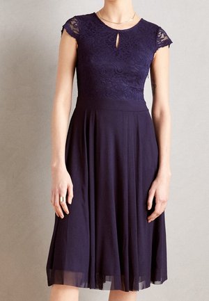 Robe bleu marine avec manches courtes en dentelle, encolure en trou de serrure, taille cintrée et jupe fluide longueur genou portée par une personne debout.