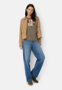 Veste en velours côtelé beige, chemise vert olive avec texte multicolore "FOREVER", jean large bleu clair et bottines vert foncé à enfiler.