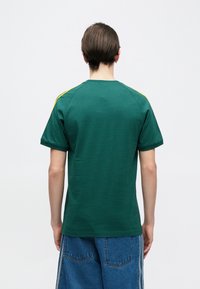 Persona ar īsiem brūniem matiem, skatoties prom, valkājot zaļu īsroku kreklu ar dzeltenām svītrām uz pleciem un zilas džinsa bikses.