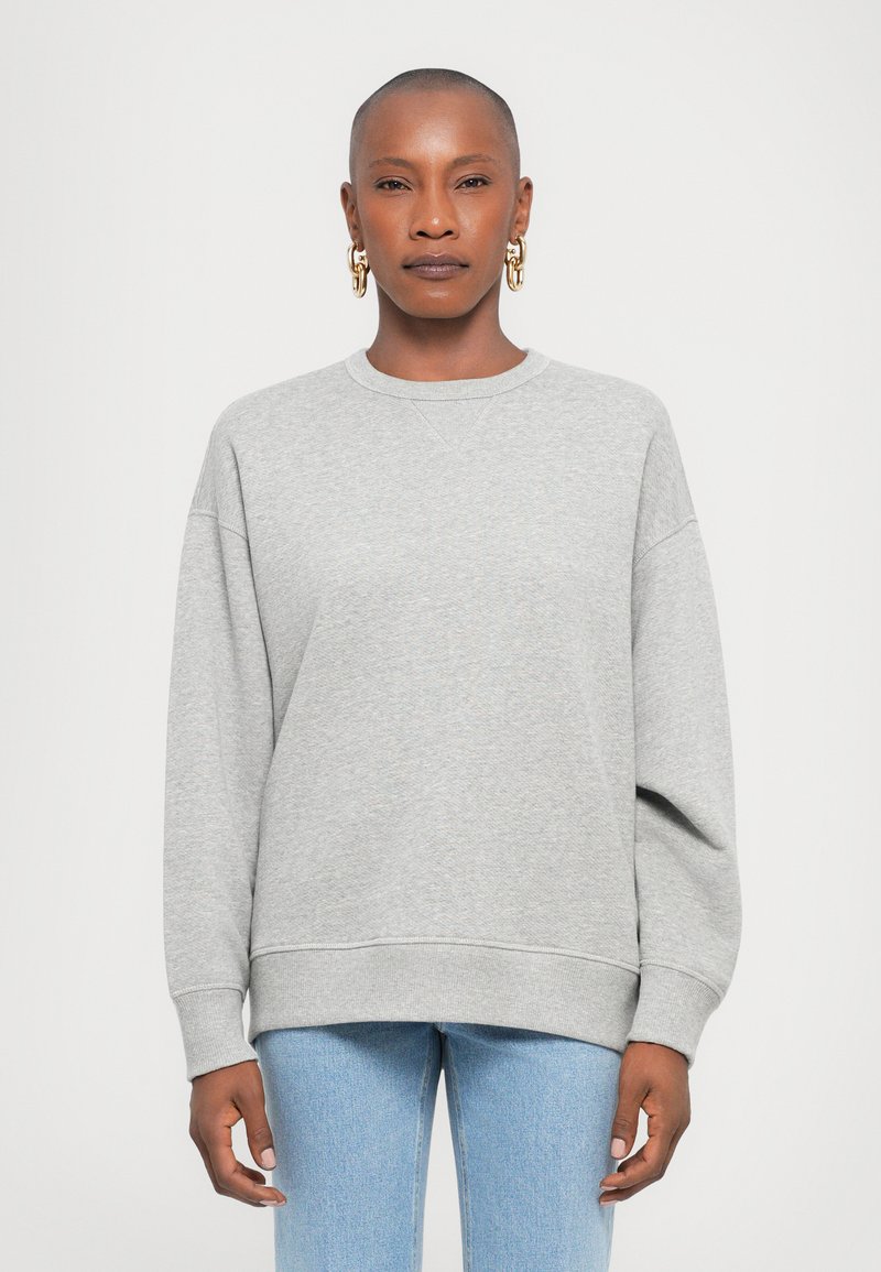 Hele hall sweatshirt ümarate kaelus, kukkunud õlad ja soonikkoes mansetid. Paaritatud helesiniste denim teksadega.