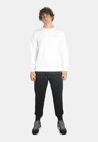 Felpa bianca a maniche lunghe con collo a girocollo, caratterizzata da un piccolo logo sul petto, abbinata a pantaloni jogger neri e scarpe da ginnastica grigie.