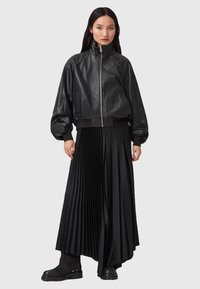 Černá kožená bomber bunda s vysokým límcem, v kombinaci s dlouhou černou plisovanou sukní a černými kotníkovými botami. Hladká textura a přiléhavý střih.