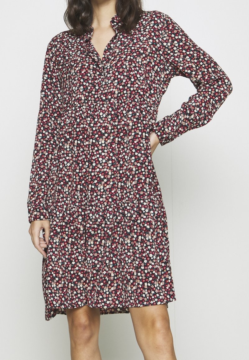Femme portant une robe à manches longues jusqu'au genou avec un petit motif floral rouge, blanc et noir, debout avec une main sur la hanche.