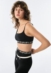 Czarny stanik sportowy z regulowanymi ramiączkami, z szerokim elastycznym paskiem z białym napisem. W połączeniu z czarnymi legginsami o wysokim stanie i sznurkiem do ściągania.