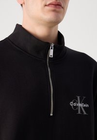 Homme portant un sweat-shirt noir à fermeture éclair quart de zip avec un zip argenté et logo Calvin Klein brodé sur la poitrine.