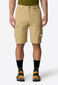 Pantaloni cargo beige in un materiale leggero, dotati di tasche frontali e laterali, dettagli cuciti e un logo sulla gamba destra.