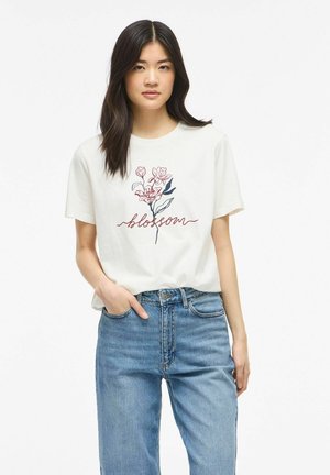 Jeune femme aux longs cheveux foncés portant un T-shirt blanc avec un motif floral et le texte « blossom », associé à un jean en denim bleu.
