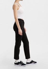 Jeans de denim negros de tiro alto, con bolsillos frontales y detalles en tono dorado, combinados con una camiseta blanca sin mangas y zapatillas negras.