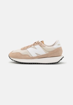 Baskets New Balance beige et blanches avec dessus en daim et mesh, logo "N" blanc, semelle coussinée et semelle extérieure noire à crampons.