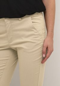 Beige tailored pants with a smooth texture, featuring side pockets and a flat front. Styled with a black top partially visible above.