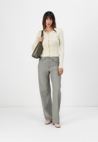 Cardigan a coste di colore crema con bottoni frontali, abbinato a pantaloni larghi grigi chiari. Il modello tiene in mano una borsa di colore verde oliva scuro e indossa scarpe chiare.
