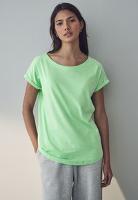 Helles grünes Baumwoll-T-Shirt mit Rundhalsausschnitt und gerollten Ärmeln, kombiniert mit grauen Sweatpants. Weiche Textur und lässige Passform.