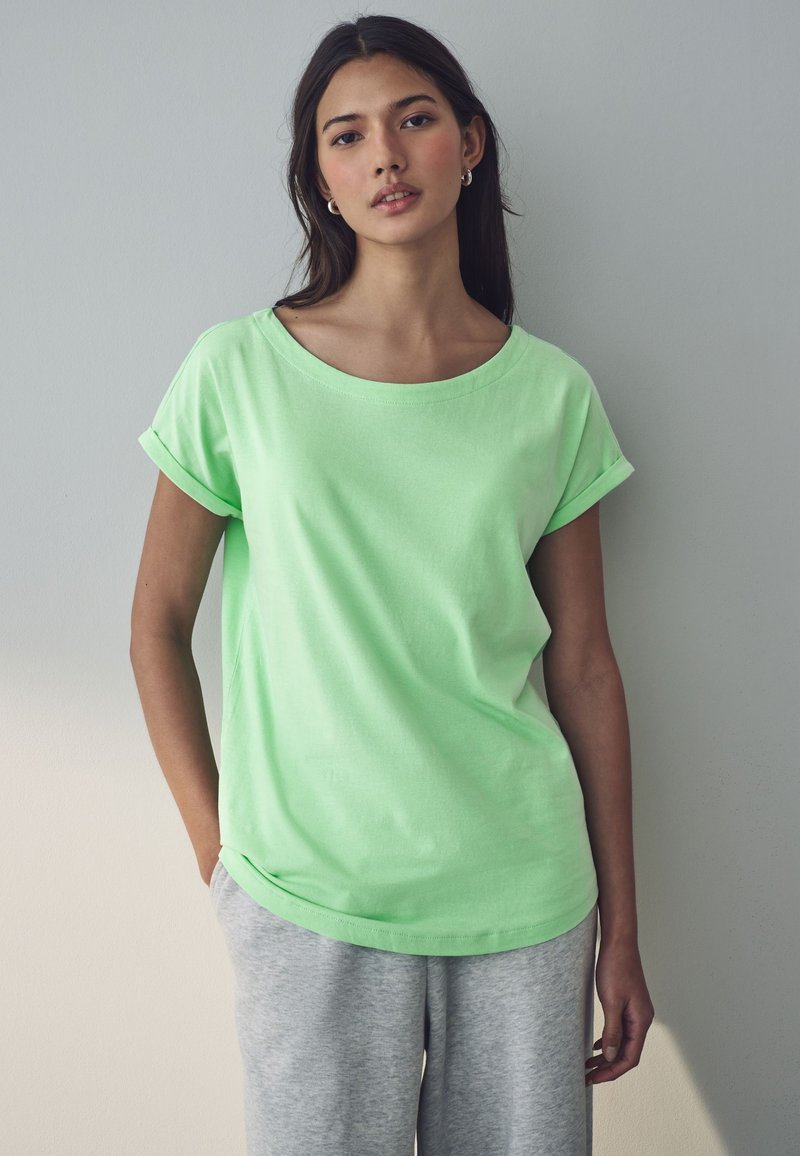 Helles grünes Baumwoll-T-Shirt mit Rundhalsausschnitt und gerollten Ärmeln, kombiniert mit grauen Sweatpants. Weiche Textur und lässige Passform.