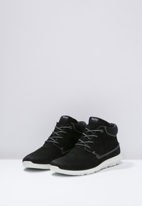 Černé semišové kotníkové boty s bílými gumovými podrážkami, které mají šedé tkaničky a prošívání. Design je jednoduchý a moderní, vhodný pro každodenní nošení.