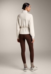 Cremefarvet pufferjakke med høj krave, indsnøret talje, parret med brune leggings og hvide sneakers, med lysegrå og lilla accenter.