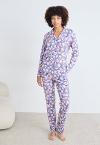 Pajama set s cvjetnim uzorkom od lagane tkanine. Košulja dugih rukava s gumbima na ljubičastoj pozadini i odgovarajuće uske hlače. Nema vidljivih metalnih dijelova.