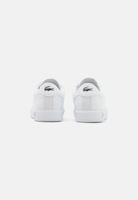 Vit sneaker med en slät läderöverkopp, vadderad krage och strukturerad gummisul. Har en svart Lacoste krokodil-logotyp på hälen.