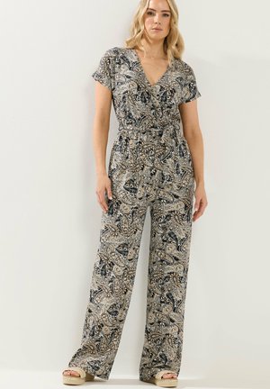 Frau trägt einen Kurzarm-Jumpsuit mit weitem Bein und Paisley-Muster, mit Wickeloptik vorne und Bindegürtel, steht vor einem einfarbigen hellen Hintergrund.