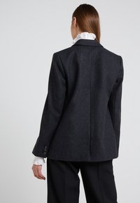 Blazer di lana grigio scuro con vestibilità su misura, rever a scollo e dettagli con tre bottoni sulle maniche. Collo bianco arricciato visibile.