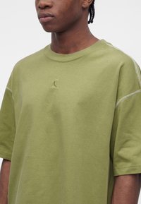 Κοντινό πλάνο ατόμου που φοράει χακί oversized μπλουζάκι με μικρό κεντητό λογότυπο Jumpman στο κέντρο του στήθους.