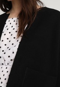 Kvinna som bär en vit smockad topp med svarta prickar och en svart öppen cardigan med en frampocket.