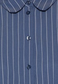Camicia chiusa con bottoni di colore blu con righe bianche verticali. Presenta un colletto arrotondato e bottoni blu lungo la parte anteriore. Tessuto morbido e testurizzato.
