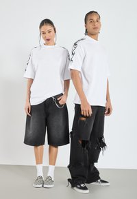 Camisetas oversized blancas con diseños de llamas negras en las mangas, combinadas con pantalones cortos de mezclilla negros y jeans negros desgastados, usados con zapatillas.