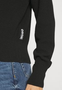 Sort ribstrikket sweater med lange ærmer og et hvidt "Hugo"-logo på siden. Kombineret med blå denimjeans med struktureret stof.
