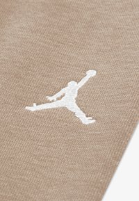 Beige stof met een wit geborduurd Jumpman-logo, centraal geplaatst, met een gestructureerd weefpatroon over het geheel.