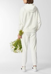 Witte hoodie en bijpassende joggingbroek gemaakt van zachte stof, met geribbelde manchetten. Model houdt een boeket bloemen vast, met zichtbare instapsneakers.