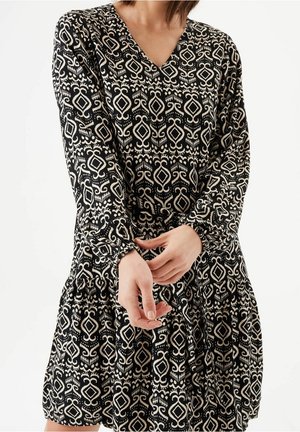 Kobieta w czarnej sukience z kremowymi geometrycznymi i spiralnymi wzorami, z długimi rękawami i dekoltem w serek, poprawiająca rękaw przy nadgarstku.