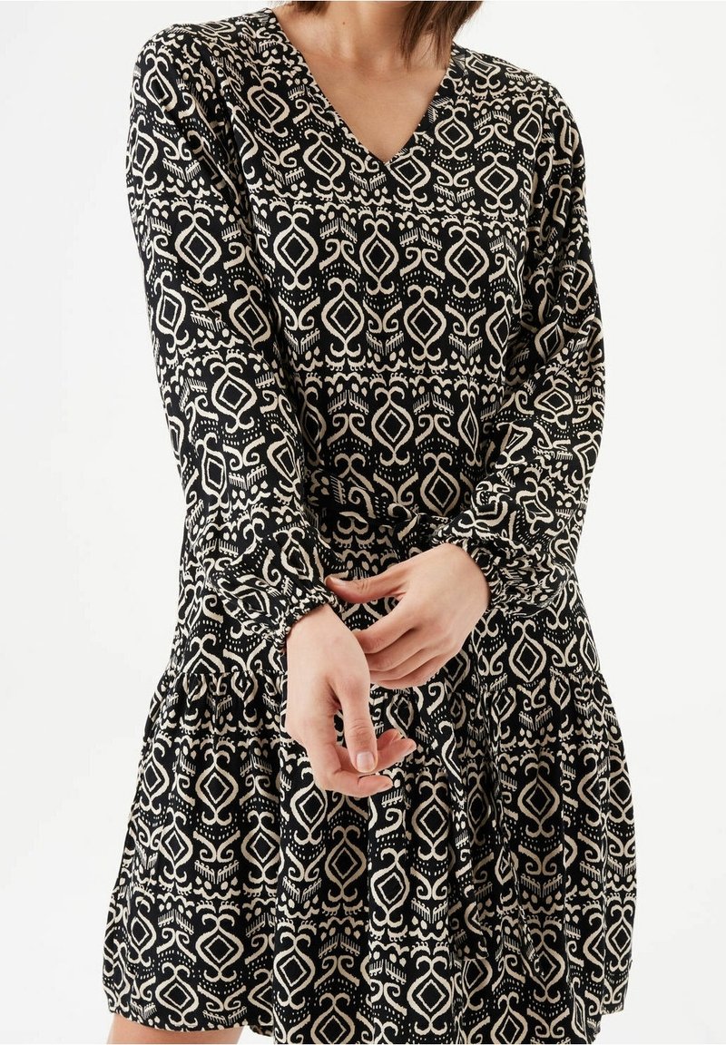 Femme portant une robe noire avec des motifs géométriques crème et des volutes, manches longues et décolleté en V, ajustant sa manche près du poignet.