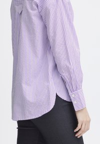 Purple and white striped button-up shirt with long sleeves, rounded hem, and buttoned cuffs, paired with dark, fitted pants.