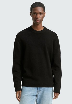 Junger schwarzer Mann trägt einen schlichten schwarzen Rundhalspullover und hellblaue Jeans, steht vor einem einfarbigen hellgrauen Hintergrund.