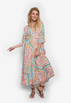 Blonde vrouw in een kleurrijke jurk met patroon tot halverwege de kuit en bruine vetersandalen, staand met één been gekruist, glimlachend tegen een witte achtergrond.