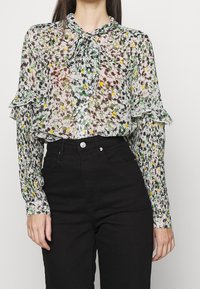 En person bär en blommönstrad, genomskinlig blus med volang ärmar, i kombination med hög-midjade svarta jeans, mot en enfärgad bakgrund.