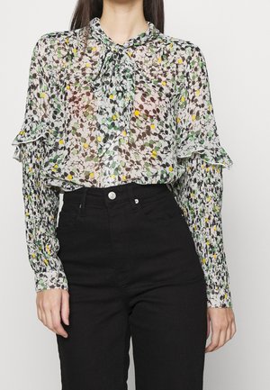 En person bærer en gennemsigtig bluse med blomsterprint og flæseærmer, parret med højtaljede sorte jeans, mod en ensfarvet baggrund.