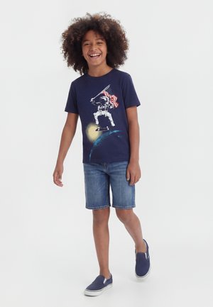 Enfant souriant aux cheveux bouclés portant un t-shirt astronaute bleu marine, un short en jean et des chaussures slip-on, debout sur un fond blanc uni.