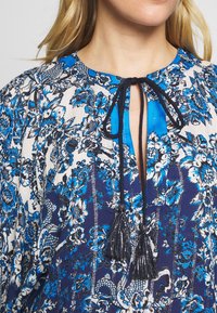 Blå och vit blommönstrad blus med rund halsringning och svarta fransar som knyts framtill, bärs av en person med blont hår.