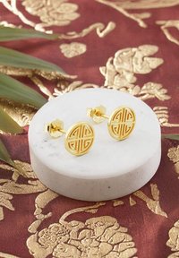 Guldtonade cirkulära örhängen med en intrikat geometrisk uttagsdesign, uppställda på en displayställ av vitt marmor. Bakgrunden har en utsmyckad tyg.