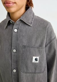 Camicia di denim grigio con colletto a punta, bottoni argentati e una tasca sul petto con un patch logo. Il tessuto ha una morbida texture vissuta.